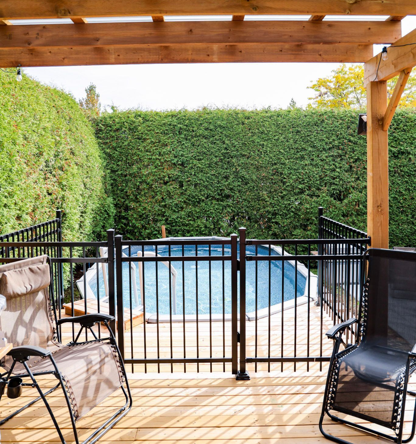 Terrasse couverte avec deux chaises donnant sur une piscine clôturée dans le jardin et une haute haie.