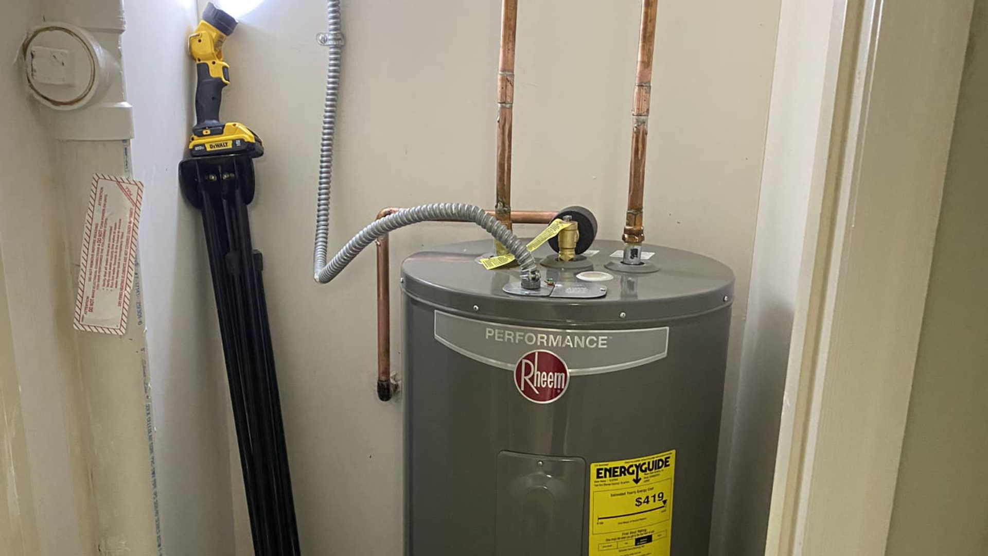 A grey Rheem water heater in a closet with copper plumbing and a yellow work light attached to the wall.