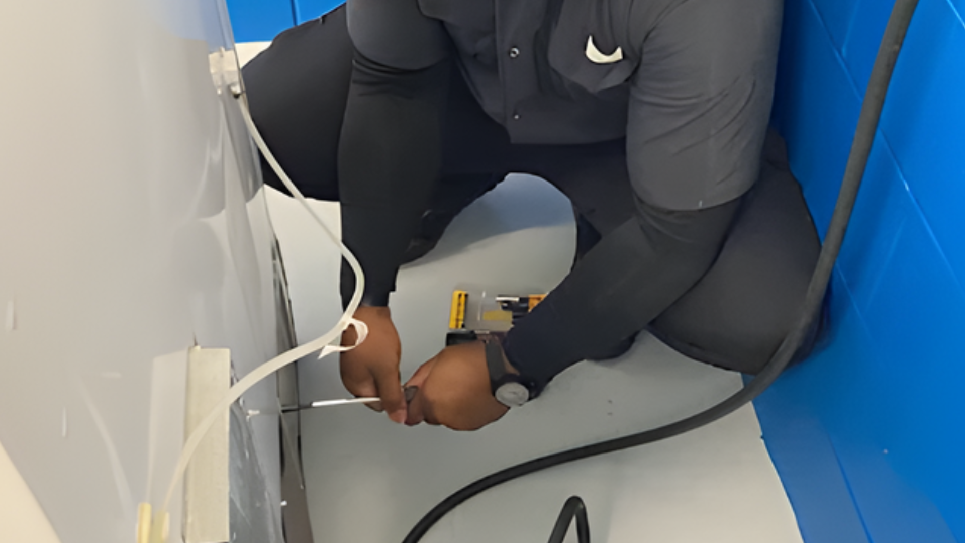 A technician wearing a dark uniform kneels on a gray floor, repairing a white appliance near a bright blue wall.