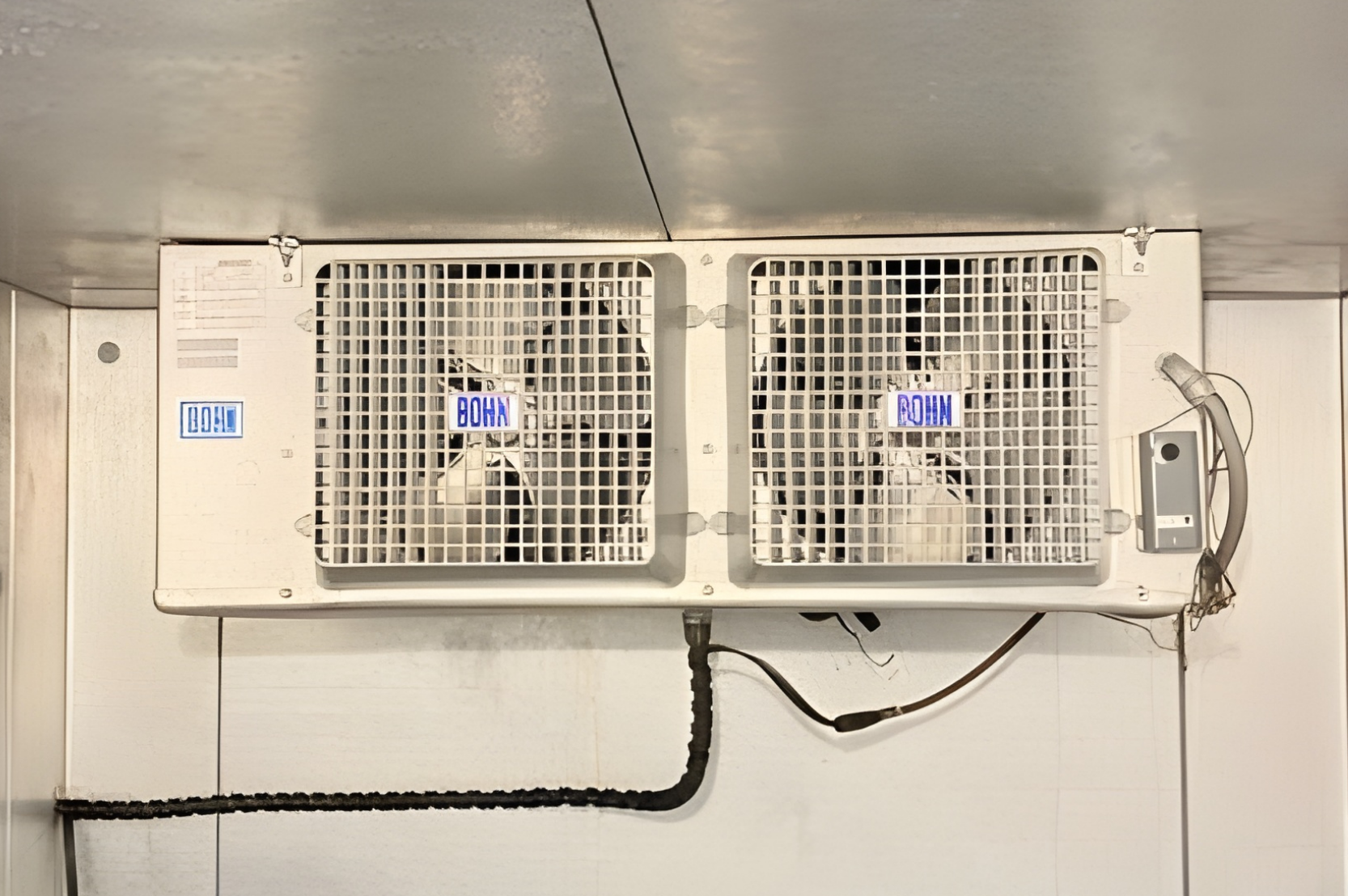 A white, dual-fan refrigeration unit mounted on a metal wall inside a cooler or freezer.