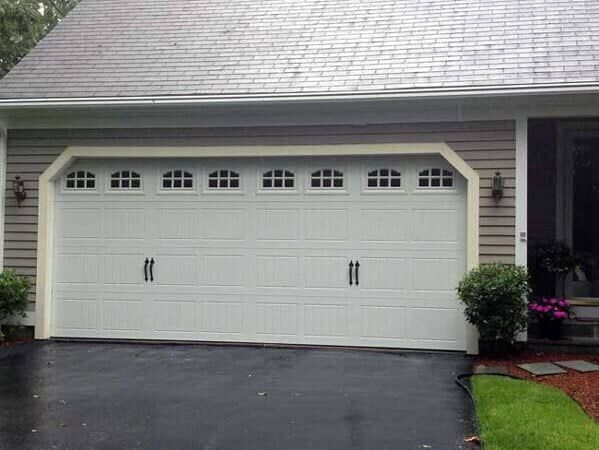 A white, two-car garage door with arched window panels and black decorative handles, set in a light gray-sided house.