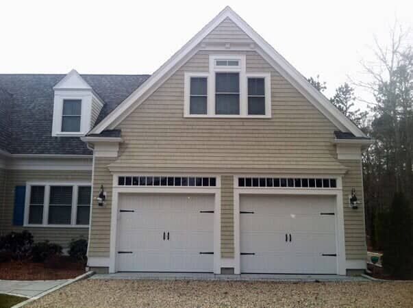 A two-car garage with white doors featuring decorative hinges and windows, attached to a tan-sided home with a gable roof.