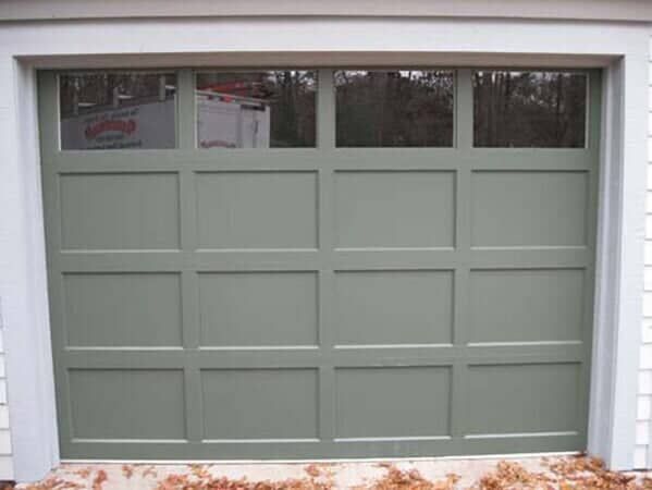 A sage green, four-panel carriage-style garage door with a row of windows at the top, installed in a home exterior.
