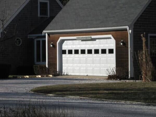 A white garage door with a row of small windows, set in a wood-sided house with a gravel driveway.