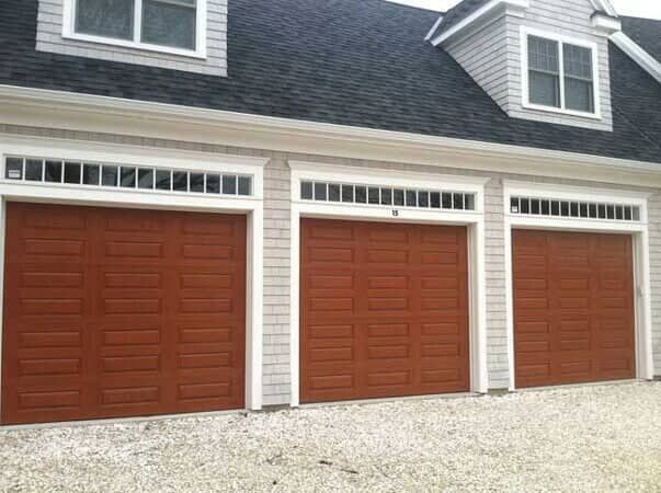 A house with three dark wood garage doors, each topped with a row of small windows, set against light gray siding.