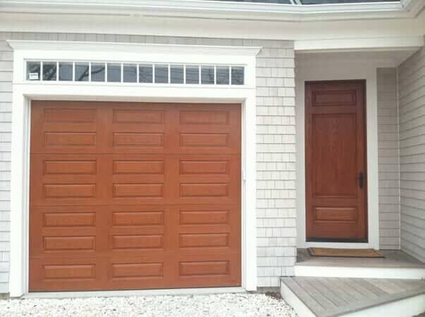 A wooden garage door with a top row of windows next to a matching wooden entry door on a house with light gray shingles.