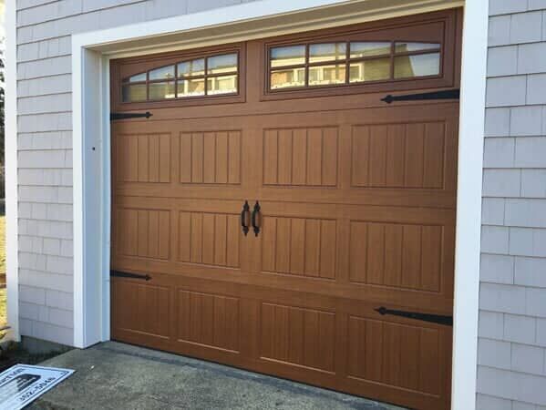A brown carriage-style garage door with windows, decorative black hinges, and central handles on a house with siding.