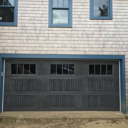 A dark gray, three-paneled garage door with windows, set against a house with light gray wood shingle siding.