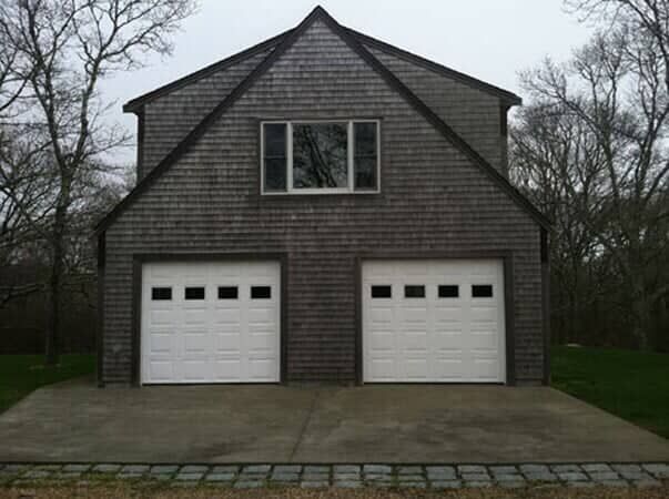A two-story, shingled garage with two white bay doors and a single window above, situated on a concrete driveway.