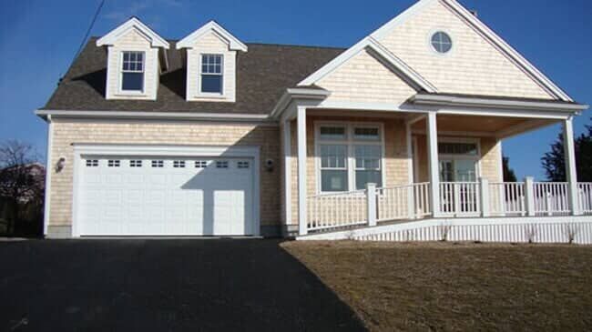 A light-colored house with cedar shingles, a white garage door, two dormer windows, and a front porch with a white railing.