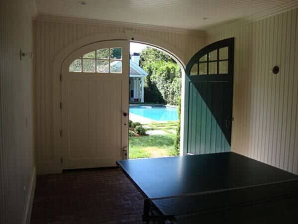 A view from inside a room with white paneled walls and a brick floor, featuring a ping pong table and open arched doors.