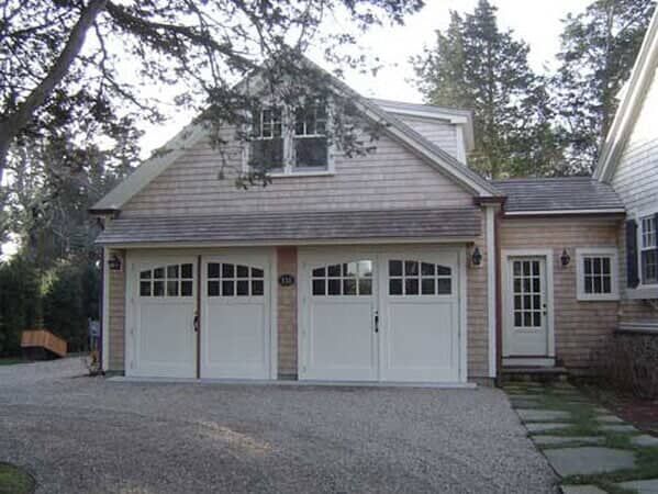 A shingled two-car garage with a loft window and a side entry door, located on a gravel driveway near trees.