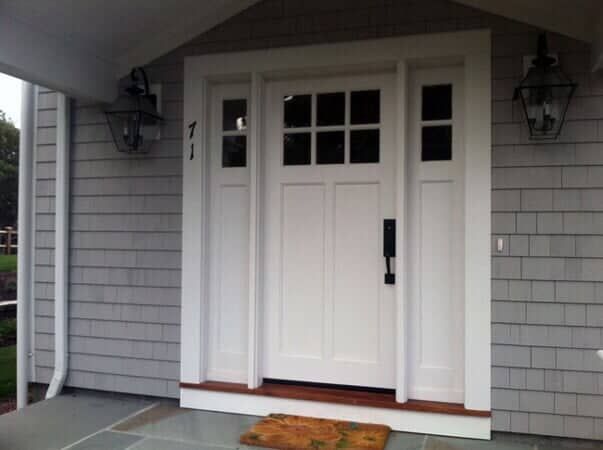 White front door with a six-pane window, sidelights, and black hardware, set in a house with light grey shingle siding.