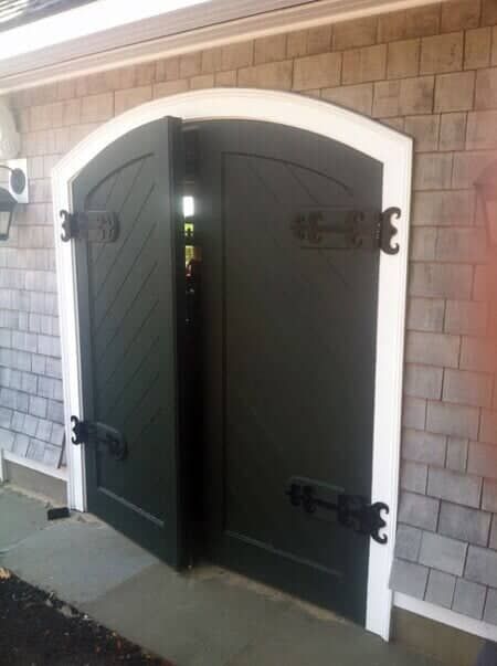 A pair of dark green, arched double doors with decorative black iron hinges, set into a shingled exterior wall.
