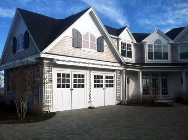 A two-story shingled home with a two-car garage, white trim, grey shutters, and a paved driveway under a blue sky.