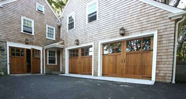 Shingle-style garage building with brown wooden carriage-style doors and a small entry door on a paved driveway.