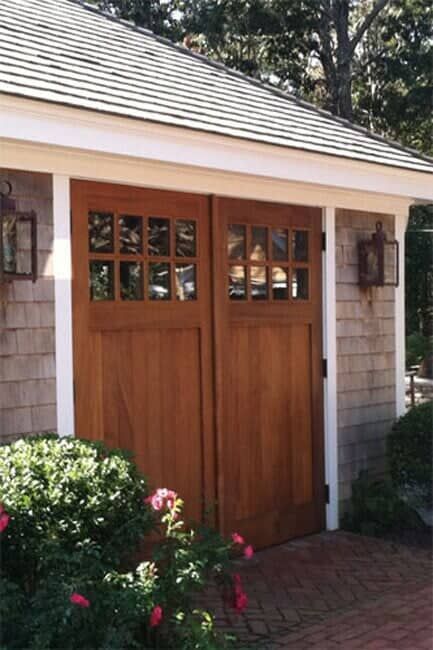 A wooden double door with a six-pane window top, set in a shingled building wall with lanterns and a brick walkway.