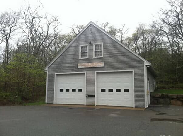A grey, two-story wooden building with two white garage doors, set against a background of trees.