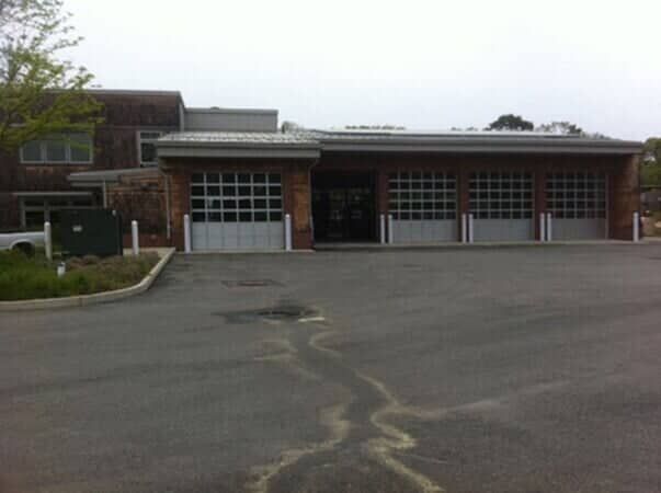 A red brick fire station building with four large, gray roll-up garage doors, set against a cloudy sky.