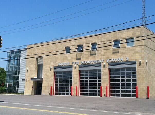 Tan stone fire department station with three glass garage doors and an adjacent modern glass stair tower.