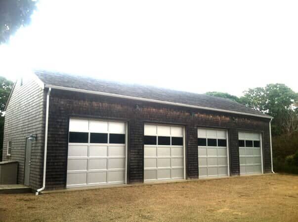 A dark shingled garage with four white multi-panel garage doors, set against a background of trees on a grassy lot.