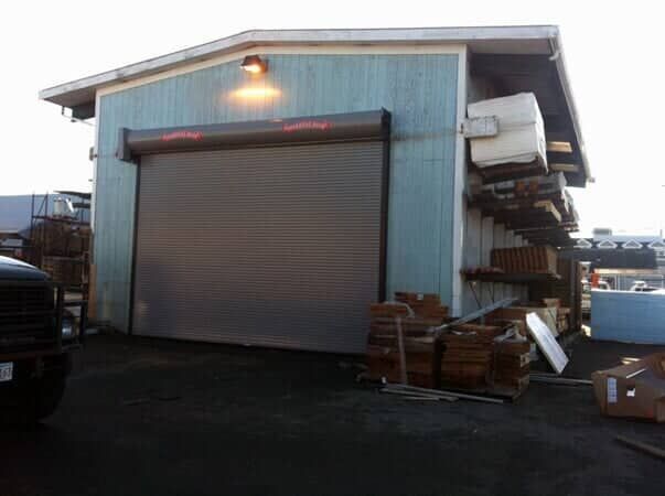 A blue warehouse exterior with a closed metal rolling door and stacks of construction materials on the side.