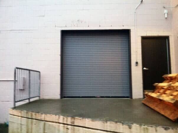 A gray roll-up industrial door beside a dark pedestrian door on a white block wall, next to a loading dock and railing.