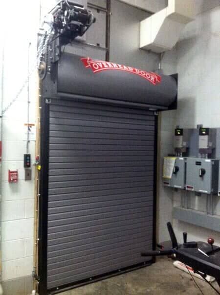 A grey metal roll-up security door with an Overhead Door logo, mounted on an interior wall with electrical boxes nearby.