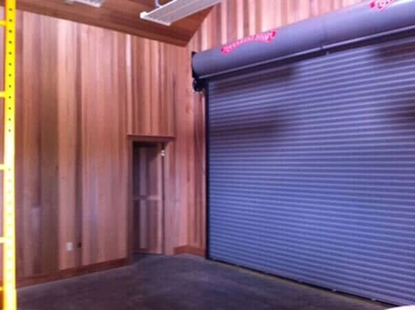 An interior view of a room with wood-paneled walls, a small door, and a large, grey industrial roll-up garage door.