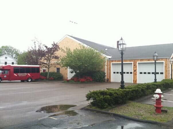 A red bus parked in front of a tan brick building with two garage bays, a fire hydrant, and a hedge in the foreground.
