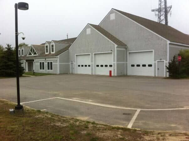A gray, multi-bay fire station with a paved parking lot in front under an overcast sky.