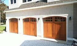 Three wooden garage doors with arched tops and glass window panels on a house with white siding and gray shingles.