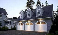 A gray, three-car garage with arched doors and three gabled dormers on the roof, viewed at dusk.