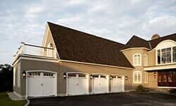 A multi-car garage with white doors, tan exterior walls, a dark shingled roof, and an upper-level balcony.