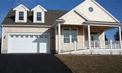 A light beige suburban house with a two-car garage, two dormer windows, and a covered porch with a wheelchair ramp.