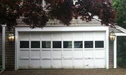 A white, multi-paneled garage door with rectangular windows, flanked by two wall-mounted lanterns under tree branches.