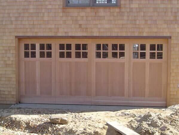 A wooden four-panel garage door with square-paned windows, installed in a house with light-colored cedar shake siding.