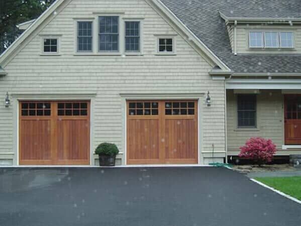 A beige, two-story house with two wooden, carriage-style garage doors, dark trim, and a small red bush in the front yard.