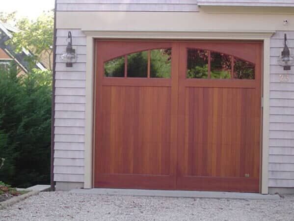 A reddish-brown wooden carriage-style garage door with two arched window panels, set in a house with light-colored siding.