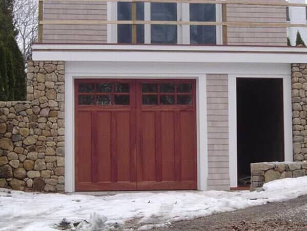 A detached two-story garage with a reddish-brown carriage-style door, stone pillars, and snow on the ground.