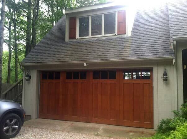 A wooden, multi-section garage door with small windows, set in a light gray exterior wall beneath a roof dormer.