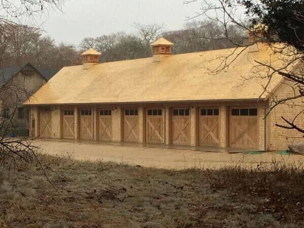 A wide, light wood-toned garage building with eight bay doors and two cupolas, situated in a wooded landscape.