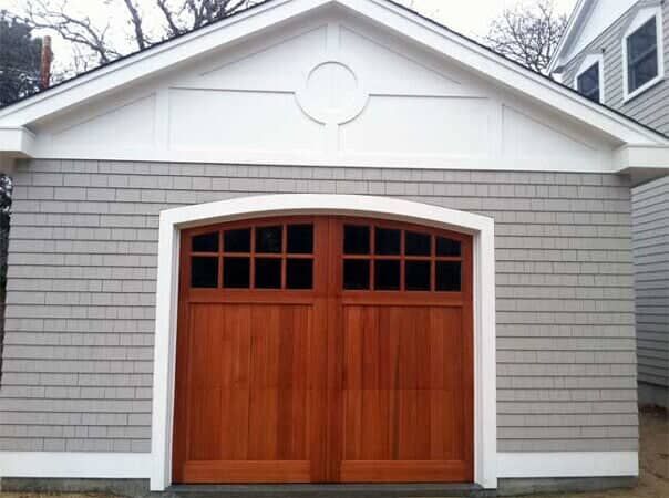 A detached garage with gray shingles, a white gabled roof, and a wooden carriage-style door with small glass panes.