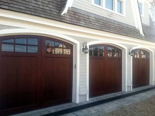 Three arched, dark wood garage doors with top window panels set in a light gray shingled house exterior.