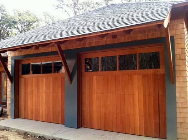 Two wooden garage doors with glass window panels set within a modern building featuring cedar shake siding.