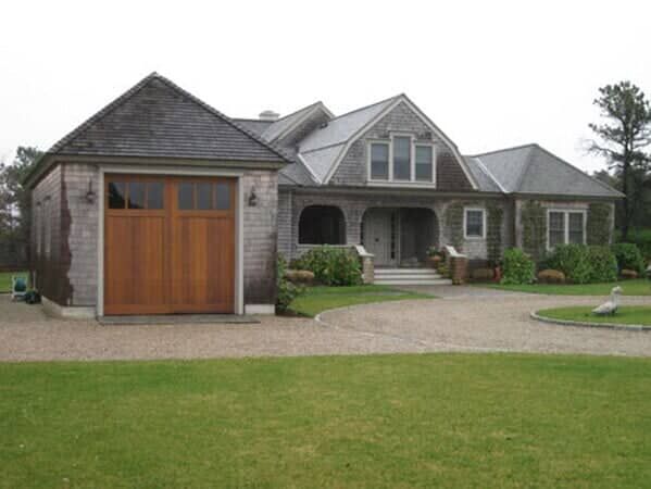 A shingle-style house with a wooden garage door and a gravel driveway set against a lawn and trees.