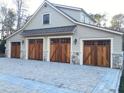 A light gray, two-story garage with three wooden carriage-style doors and stone accents on a stone-paved driveway.
