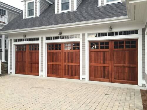Three wooden carriage-style garage doors with window inserts set in a grey-sided house with a paved driveway.