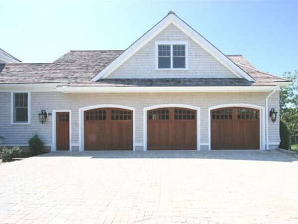 A light-gray shingled house with three wooden garage doors and a stone driveway under a clear blue sky.