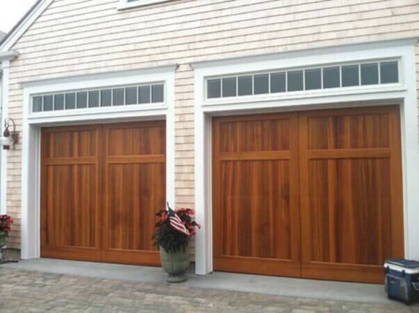 Two wooden garage doors with glass-paned transoms above, installed in a shingled building with a potted plant outside.
